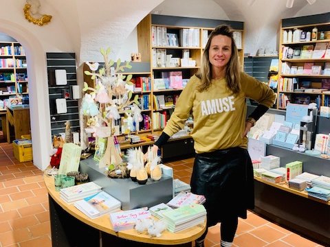 Eine lächelnde Frau in einem charmanten Laden mit Büchern, Geschenken und Gewölbedecken, an einem Präsentationstisch.