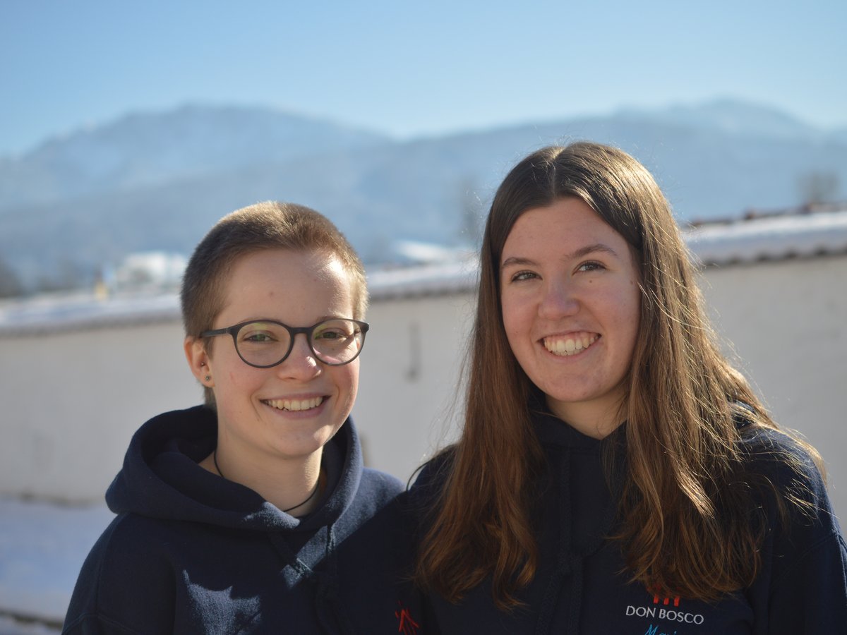 Zwei lächelnde junge Frauen stehen draußen im Winter, mit schneebedeckten Bergen und blauem Himmel im Hintergrund.
