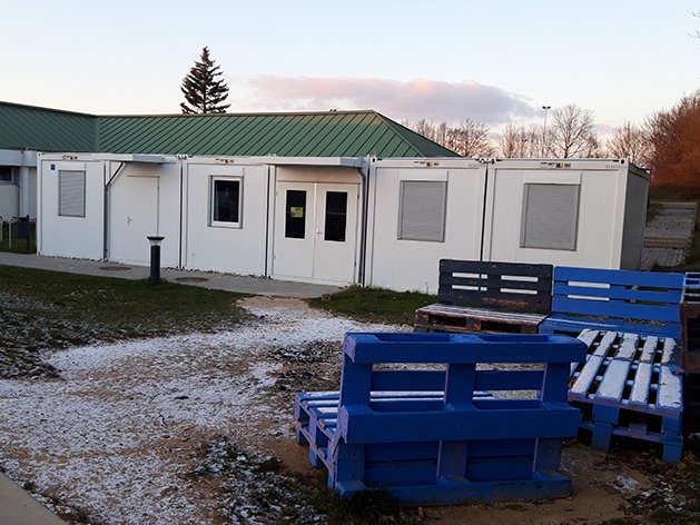 Weiße Modulgebäude mit grünem Dach und blau-dunklen Palettenbänken, leicht mit Schnee bedeckt an einem Wintertag.