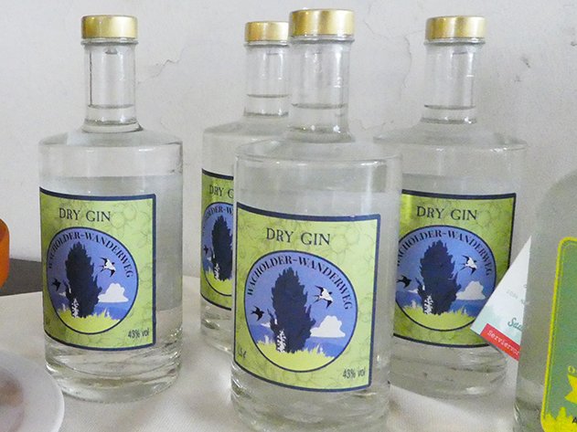 Mehrere klare Flaschen "Dry Gin Wacholder-Wanderweg" mit markanten grün-blauen Etiketten, die einen Baum und Vögel zeigen.