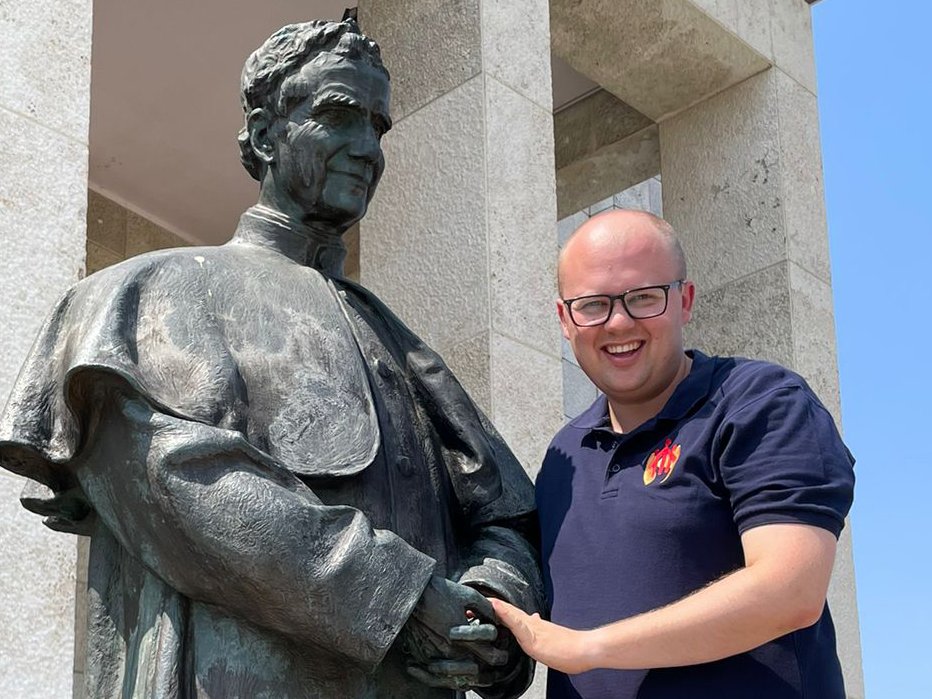 Ein lächelnder Mann mit Brille steht neben einer Bronzestatue einer bekleideten Figur und berührt sanft ihre Hand.
