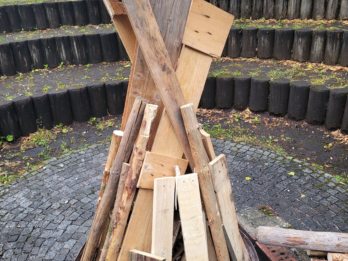 Großer Tipi-förmiger Holzstapel in einer Metall-Feuerschale auf Kopfsteinpflaster, mit gestuften Holzsitzen und Grün im Hintergrund.