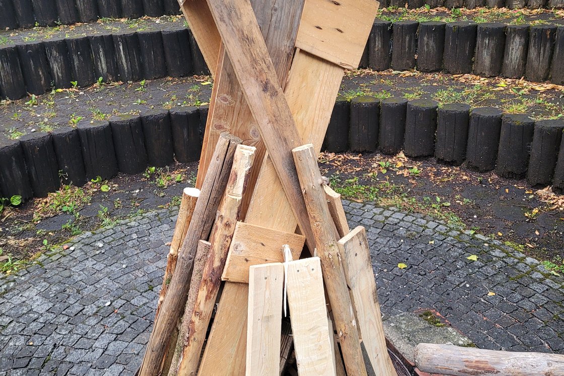 Großer Tipi-förmiger Holzstapel in einer Metall-Feuerschale auf Kopfsteinpflaster, mit gestuften Holzsitzen und Grün im Hintergrund.