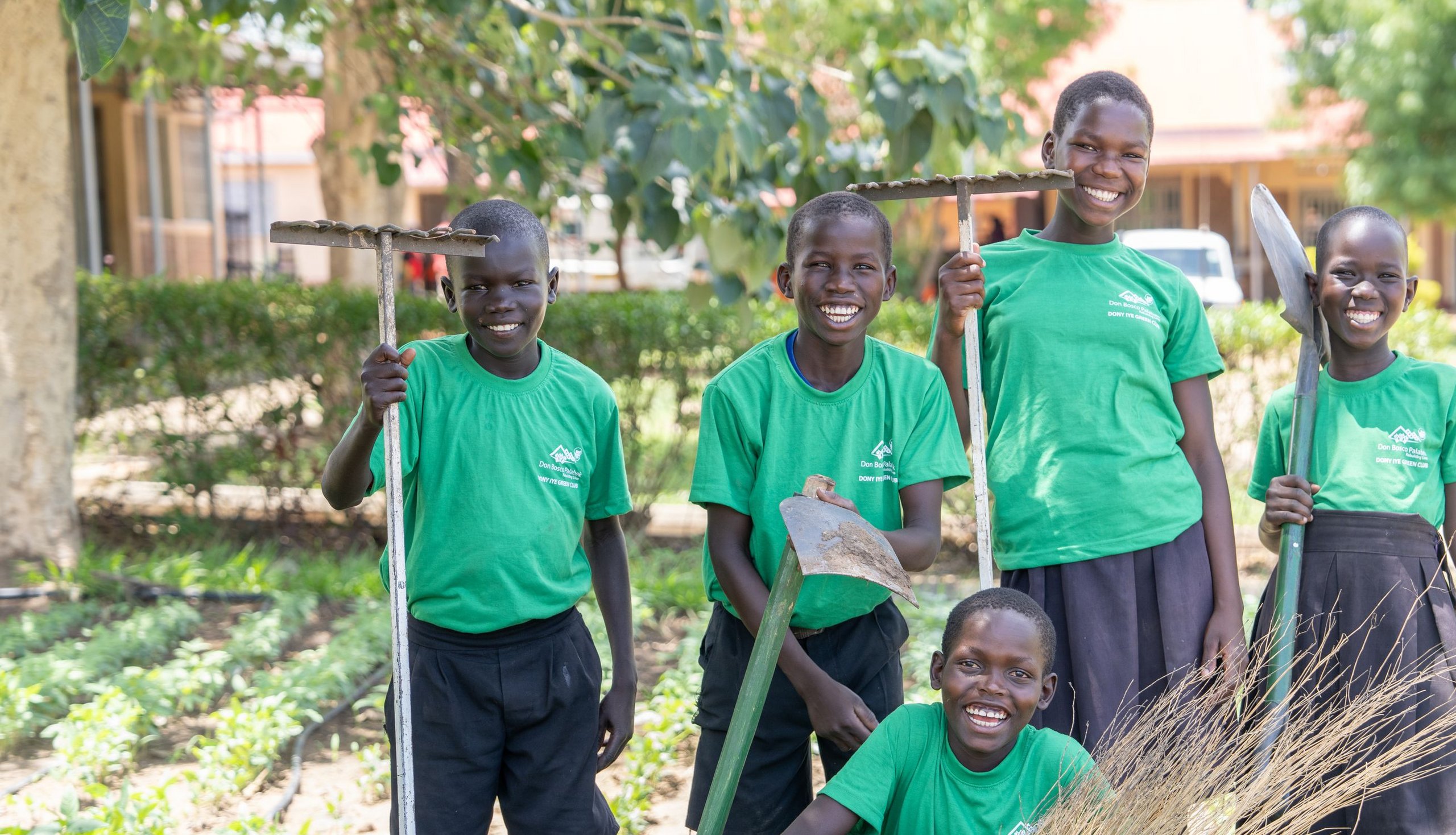 Fünf lächelnde Jugendliche in grünen Shirts halten Gartengeräte in einem üppigen Garten mit Pflanzenreihen.