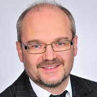 Porträt eines lächelnden Mannes mittleren Alters mit Brille, Ziegenbart und grauem Haar, im Nadelstreifenanzug.