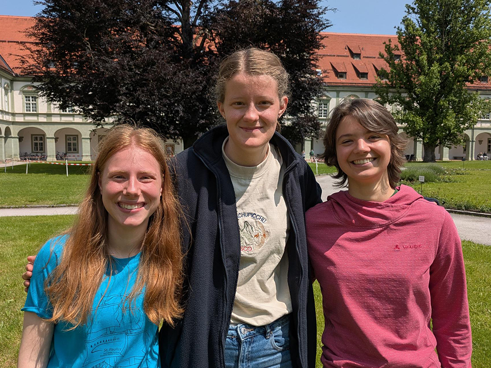 Drei lächelnde junge Frauen stehen an einem sonnigen Tag im Freien vor einem historischen Gebäude mit Bögen und Bäumen.