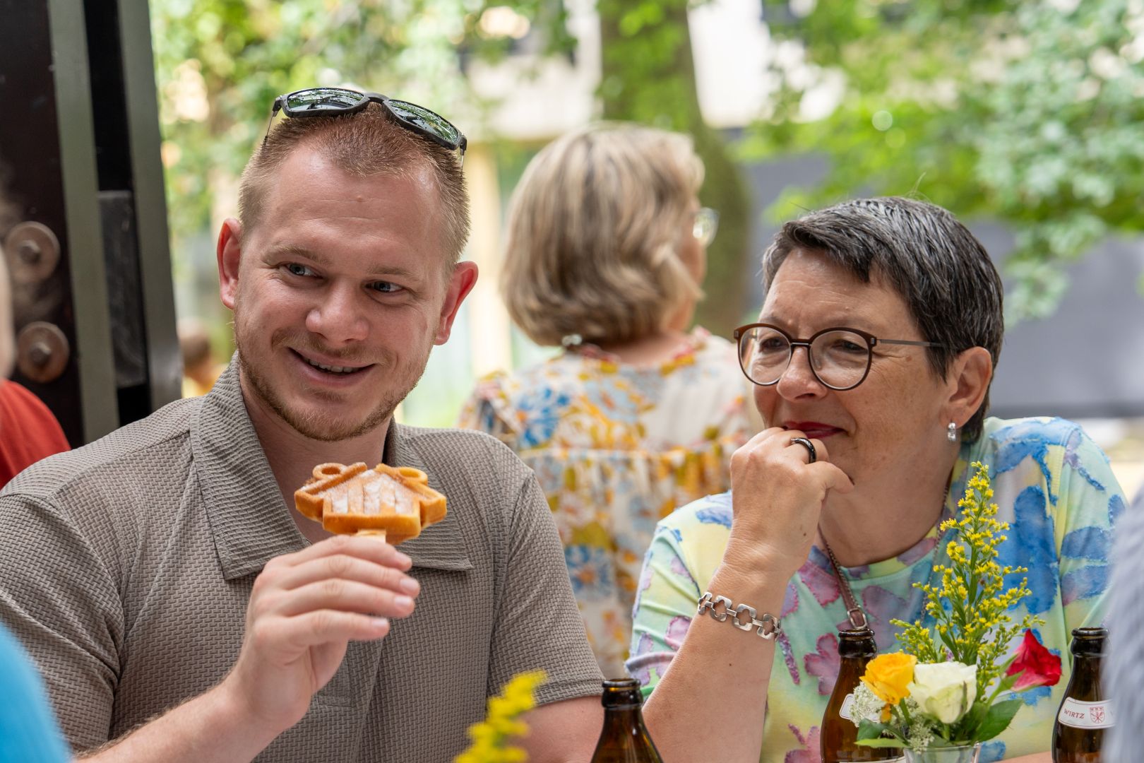 Ein lächelnder Mann hält ein hausförmiges Gebäck, daneben eine lächelnde Frau mit Brille an einem Tisch im Freien.