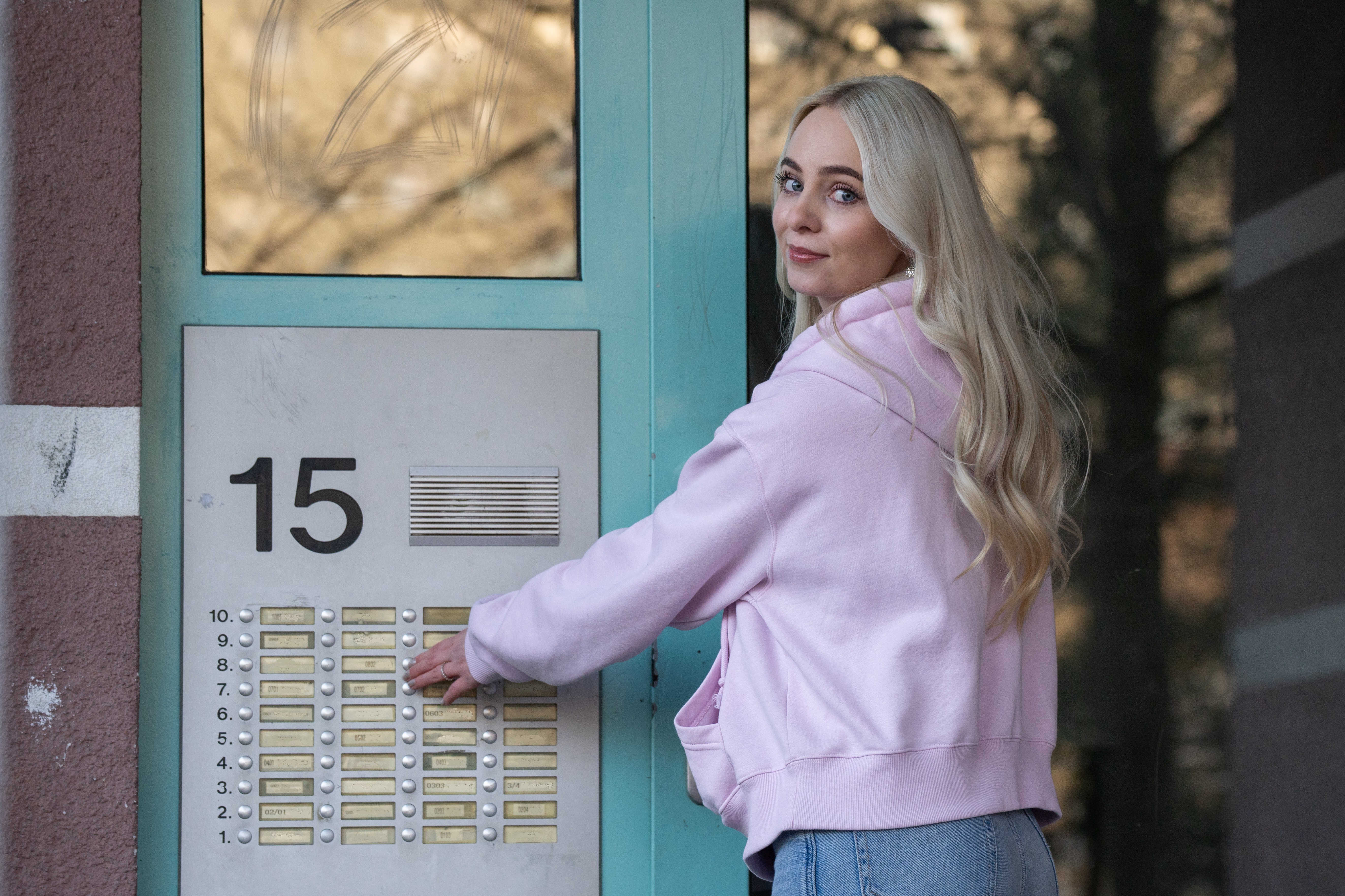 Blonde Frau in pinkfarbenem Hoodie drückt Klingelknopf an Gegensprechanlage mit Nr. 15, blickt zurück.