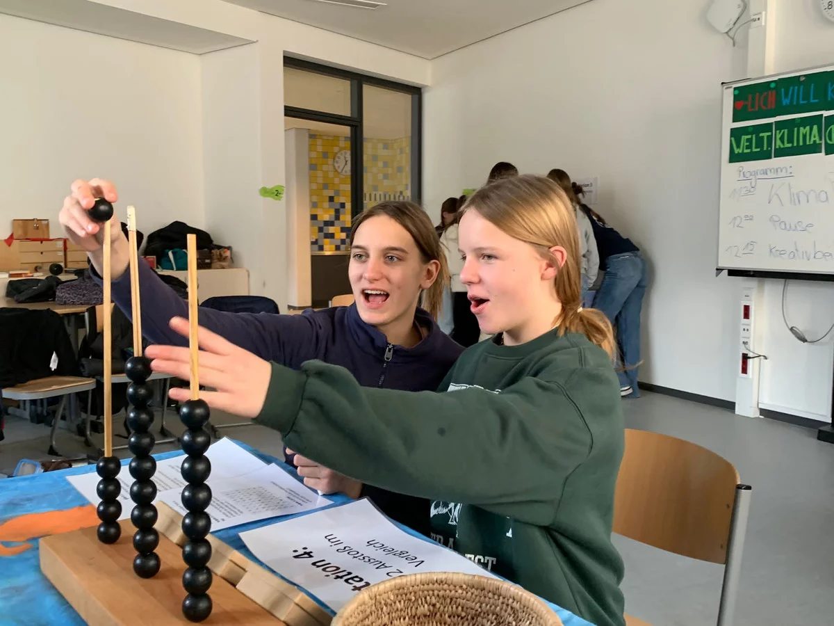 Zwei Mädchen beschäftigen sich enthusiastisch mit einem Zählgerät aus Perlen in einem Klassenzimmer, wohl bei einer Klima-Aktivität.