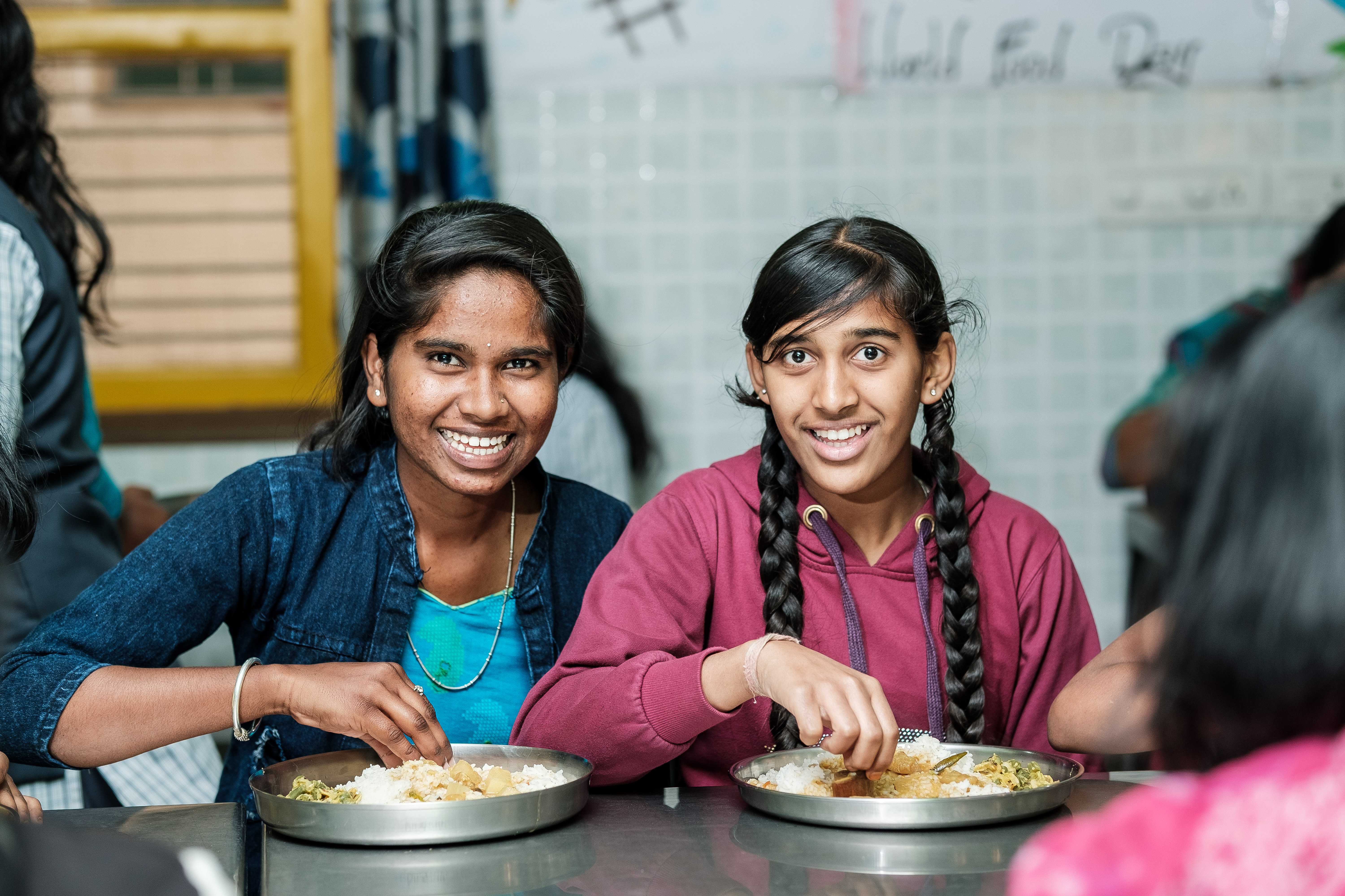 Zwei lächelnde junge Frauen essen mit ihren Händen Reis und Curry von Metalltellern in einem Gemeinschaftsbereich.