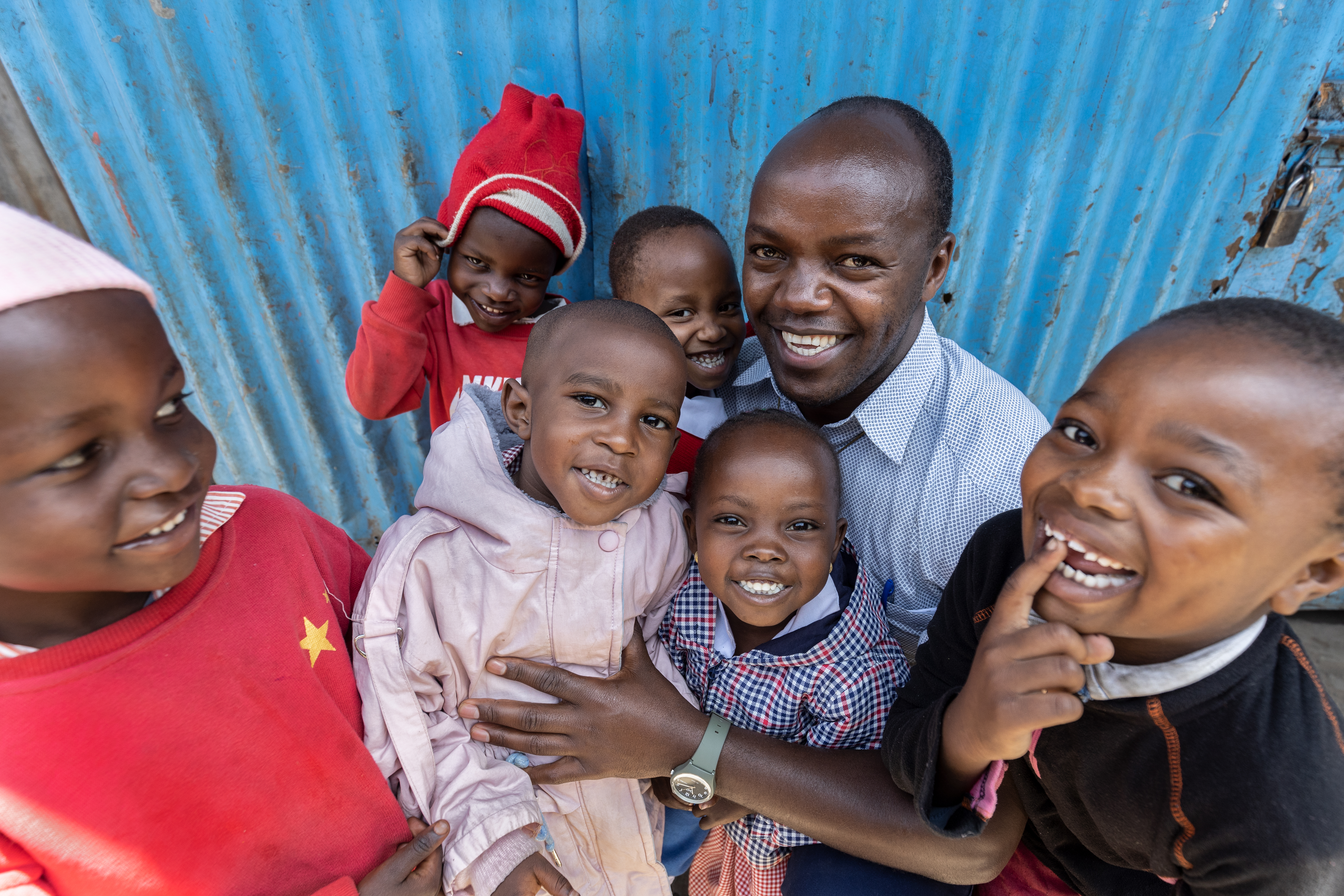 Ein lächelnder Mann umarmt mehrere lachende Kinder vor einer blauen Wellblechwand.