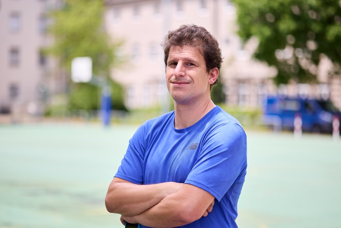 Ein Mann steht vor einem Sportplatz und lächelt.