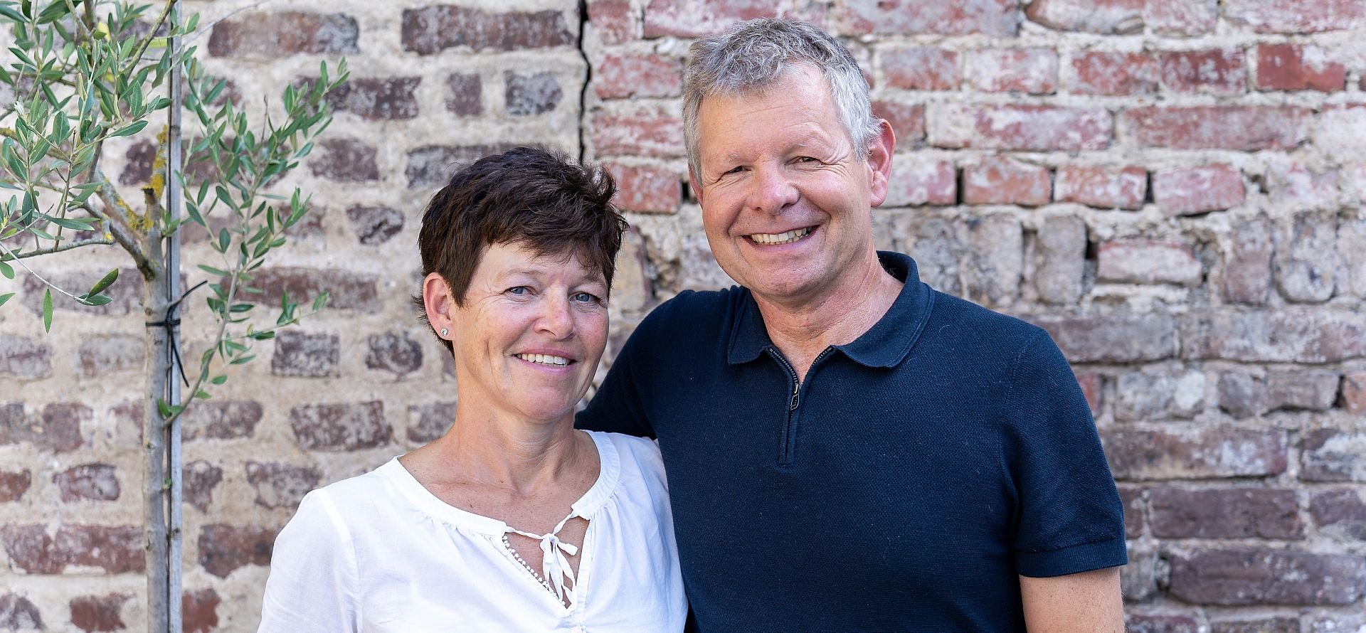 Lächelndes Paar, eine Frau mit kurzen braunen Haaren und ein Mann mit grauen Haaren, vor einer Ziegelmauer.