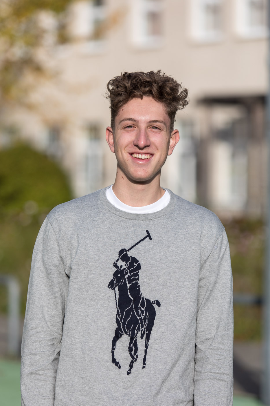 Ein lächelnder junger Mann mit lockigem Haar, der ein graues Sweatshirt mit großem Ralph Lauren Polo-Logo trägt. Im Freien.