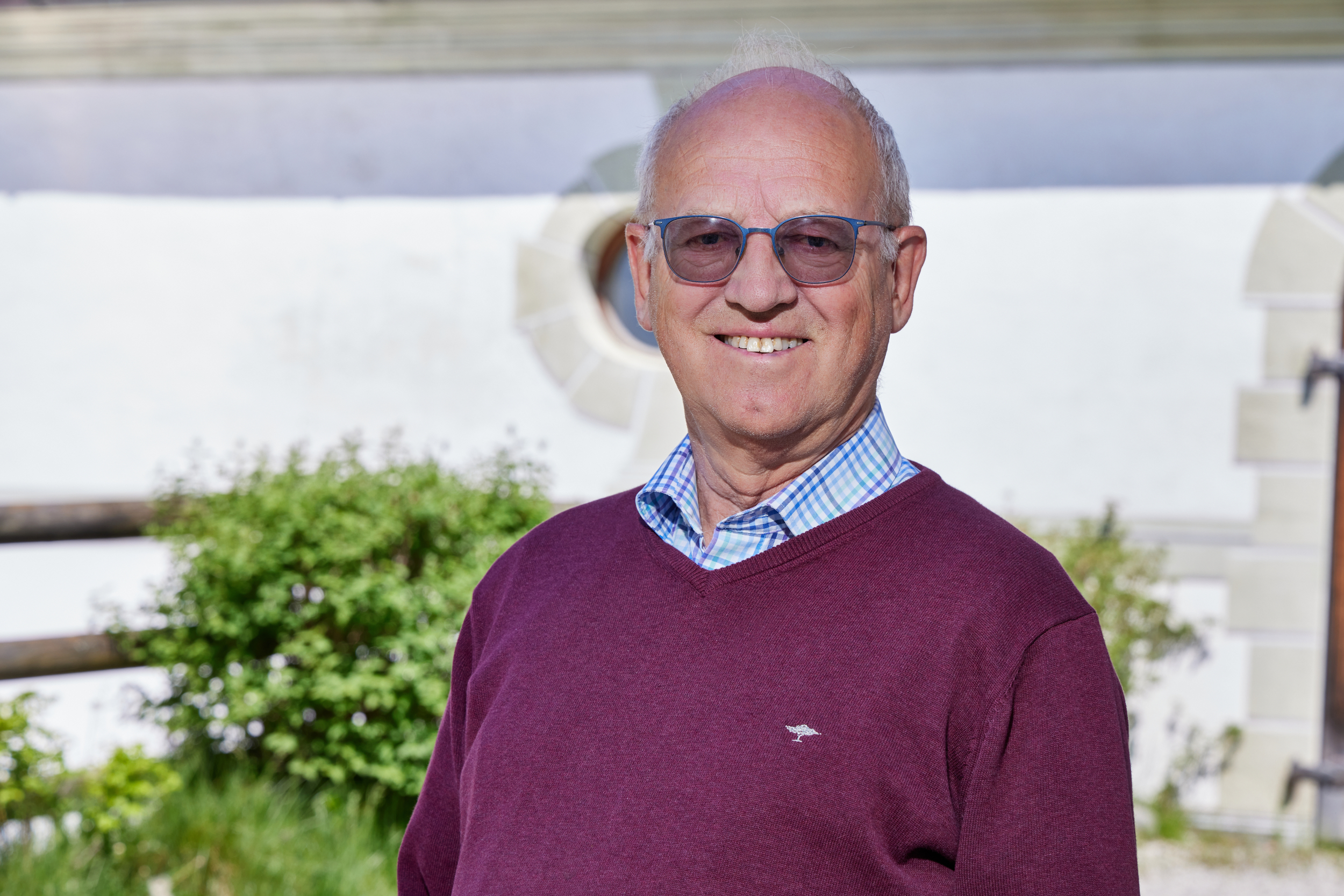 Porträt eines lächelnden älteren Mannes mit Brille und dunkelrotem Pullover vor verschwommenem Hintergrund.