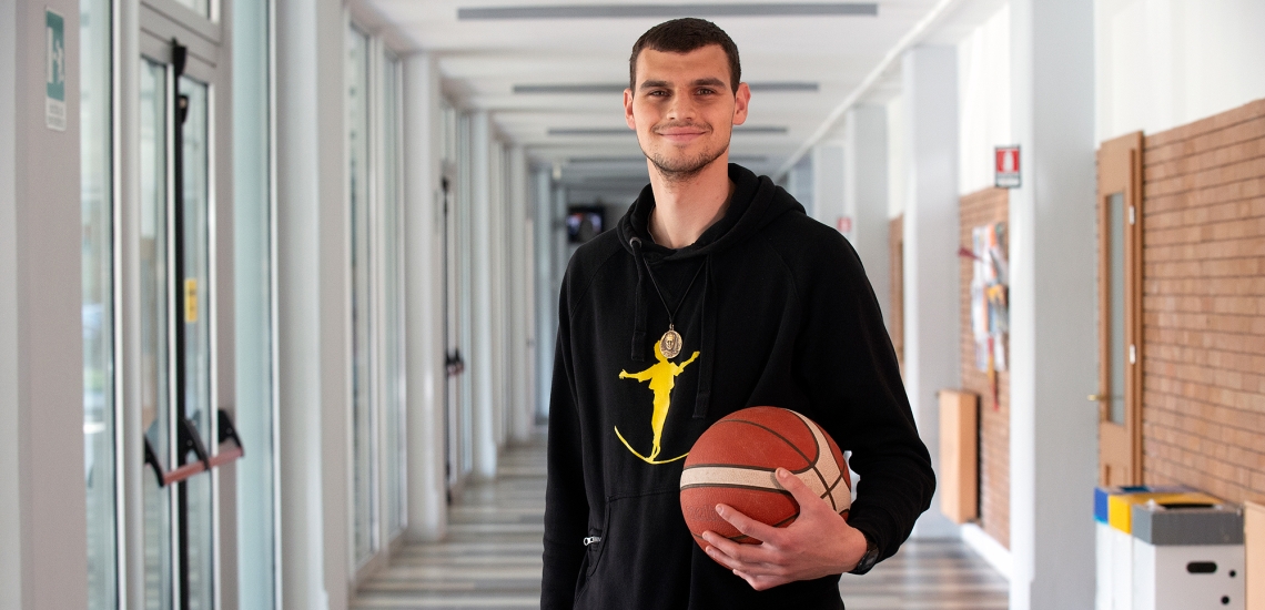 Lächelnder junger Mann in einem schwarzen Kapuzenpullover, der einen Basketball in einem modernen Flur hält.