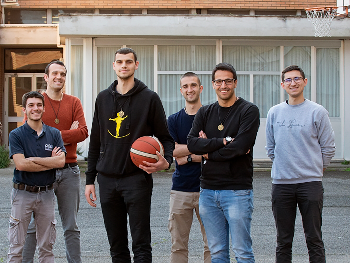 Sechs lächelnde junge Männer, einer hält einen Basketball, stehen draußen vor einem Gebäude mit Basketballkorb.