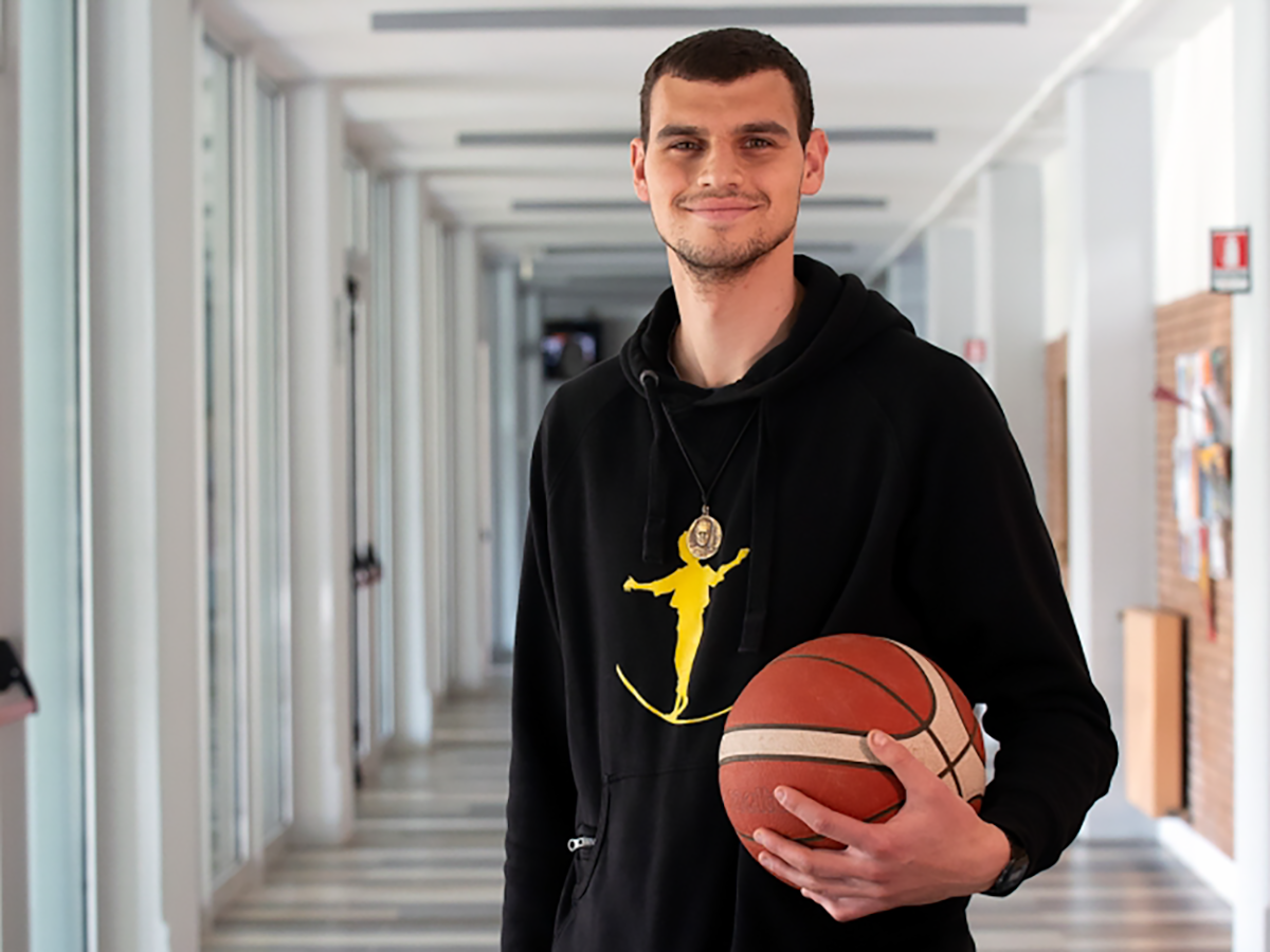 Ein lächelnder junger Mann in einem schwarzen Kapuzenpullover mit gelbem Motiv hält einen Basketball in einem hellen Flur.