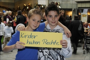 Zwei lächelnde Jungen halten ein gelbes Schild mit der Aufschrift „Kinder haben Rechte“, Arm in Arm in einem öffentlichen Raum.
