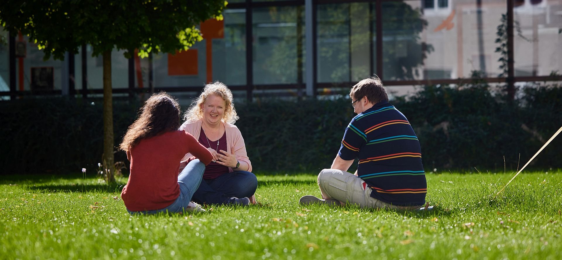 Drei Personen sitzen an einem sonnigen Tag draußen auf einer Wiese, eine Frau lächelt eine andere während eines Gesprächs an.