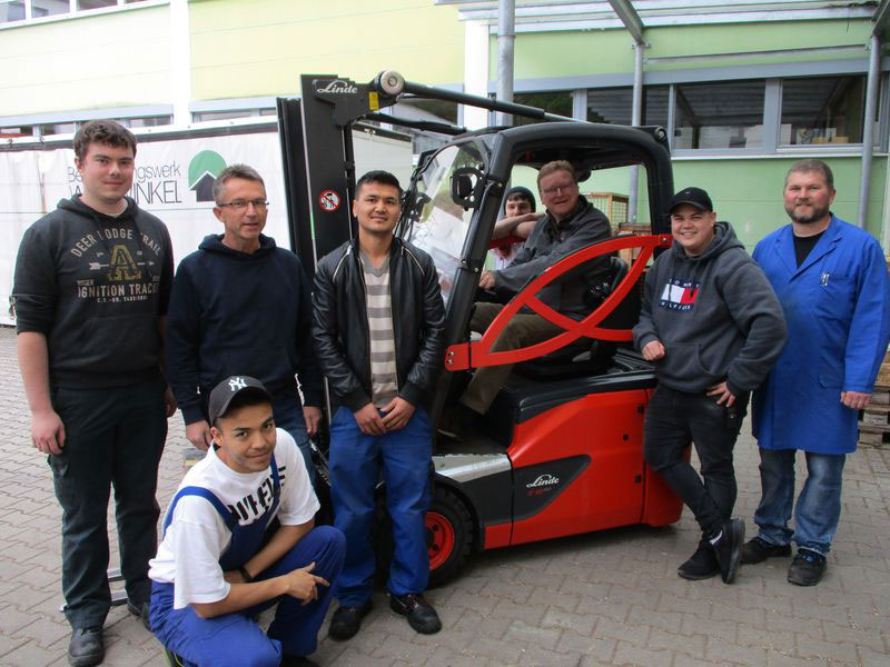 Gruppe von acht Männern, darunter Arbeiter und Studenten, lächelnd um einen roten Linde Gabelstapler im Freien.