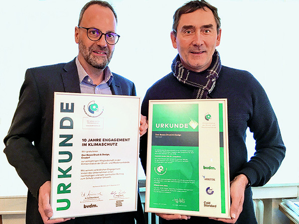 Zwei Männer präsentieren Urkunden für 10 Jahre Engagement im Klimaschutz für ihre Druckerei.