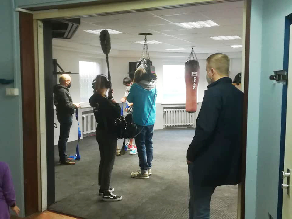 Eine Filmcrew baut in einem Fitnessstudio mit Boxsäcken auf. Ein Kameramann, Tontechniker und Beobachter sind anwesend.