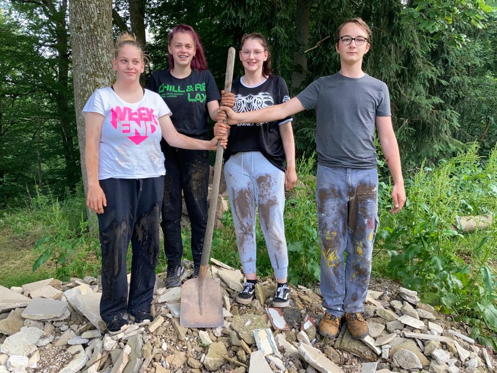 Vier Jugendliche stehen mit einer Schaufel auf einem Schutthaufen, ihre Kleidung ist schmutzig von der Arbeit.