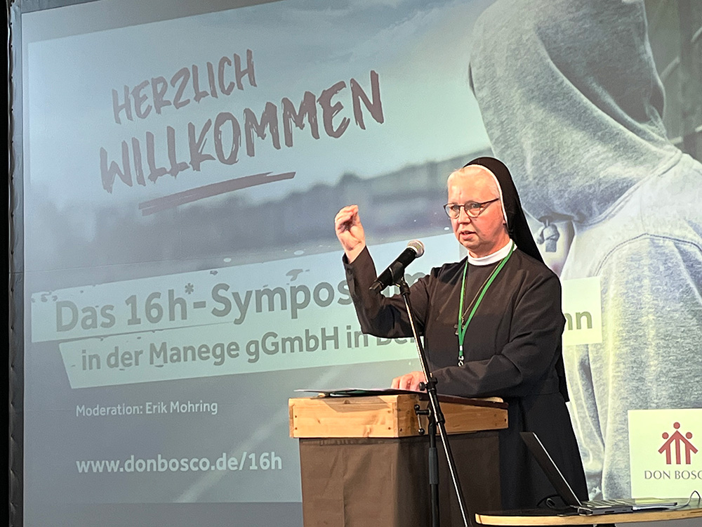 Nonne spricht auf einem Symposium, im Hintergrund Projektion mit „Herzlich Willkommen“ und Don Bosco Logo.