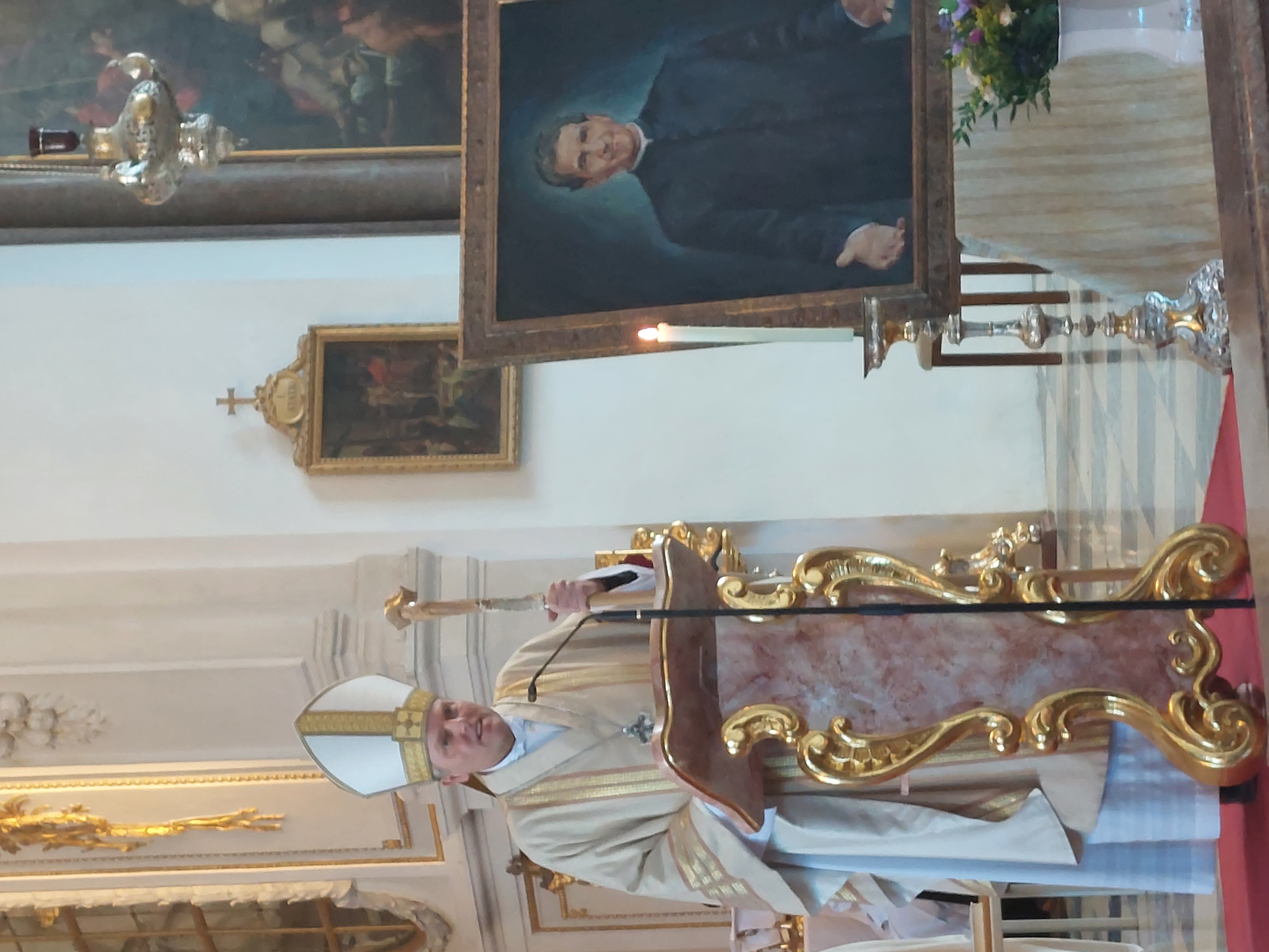 Ein Bischof mit Mitra und Krummstab spricht von einem goldenen Lesepult in einer Kirche, neben einem Porträt eines Priesters.