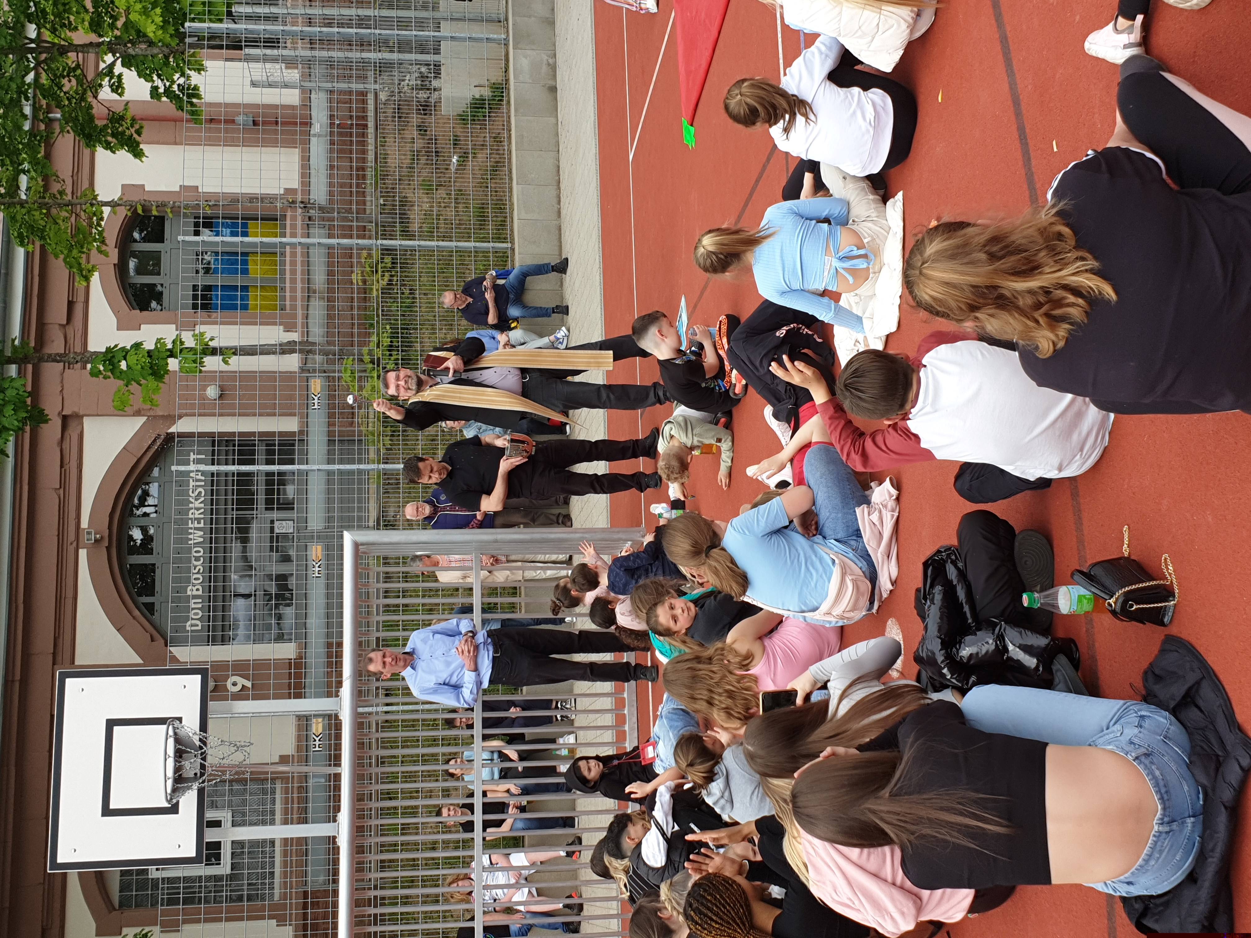 Priester segnet eine Gruppe von Jugendlichen und Kindern auf einem Sportplatz vor der Don Bosco Werkstatt mit einer Schöpfkelle.