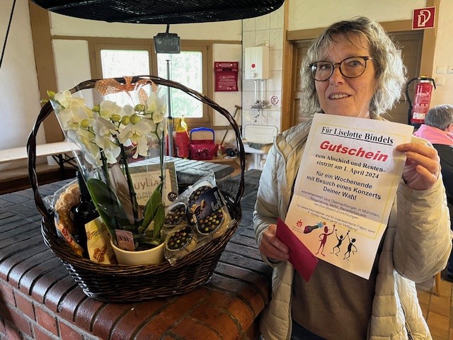 Eine lächelnde Frau hält einen Renteneintritts-Gutschein neben einem Geschenkkorb mit Orchidee und weiteren Präsenten.