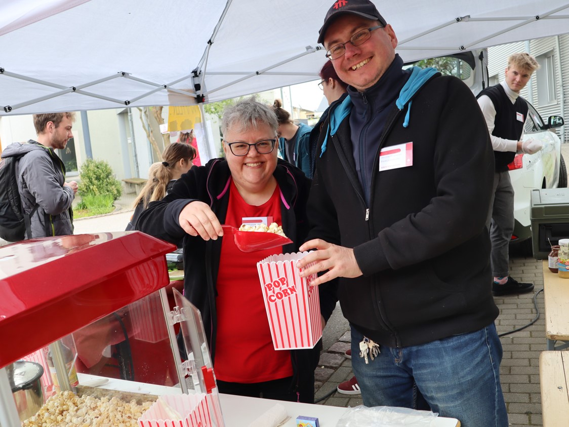 Zwei lächelnde Personen, eine Frau und ein Mann, servieren Popcorn aus einer Maschine unter einem weißen Zelt bei einer Veranstaltung.