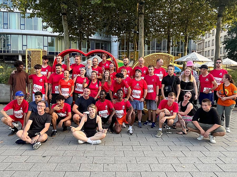 Eine große, vielfältige Gruppe von Menschen, viele in roten Shirts mit Nummern, posiert nach einem Event vor einem roten Herz.