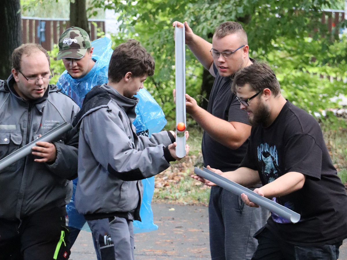 Fünf Männer arbeiten bei einer Outdoor-Teamaktivität zusammen und leiten einen orangefarbenen Ball durch eine Rinne aus grauen Rohrabschnitten.