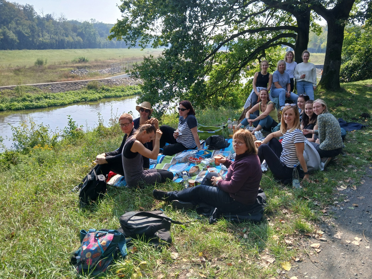 Eine große Gruppe Frauen genießt ein sonniges Picknick am Fluss, auf Gras unter Bäumen sitzend.