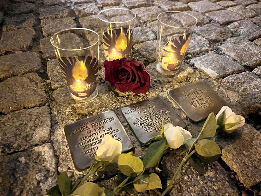 Drei Stolpersteine mit Gedenkkerzen und Rosen auf Kopfsteinpflaster bei Nacht, zum Gedenken an Holocaust-Opfer.