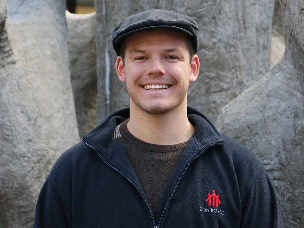 Porträt eines lächelnden Mannes mit dunkler Schiebermütze und Fleecejacke mit „Don Bosco“-Logo.