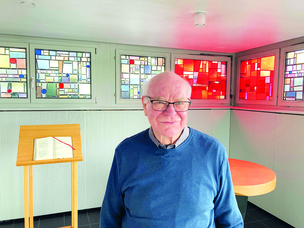 Älterer Mann mit Brille lächelt in einem modernen Raum mit abstrakten Buntglasfenstern und einem Lesepult.