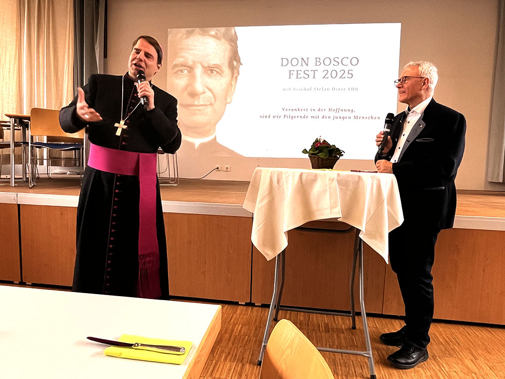 Ein Bischof und ein Mann im Anzug sprechen auf einer Bühne. Im Hintergrund: eine Projektion von Don Bosco und "Don Bosco Fest 2025".