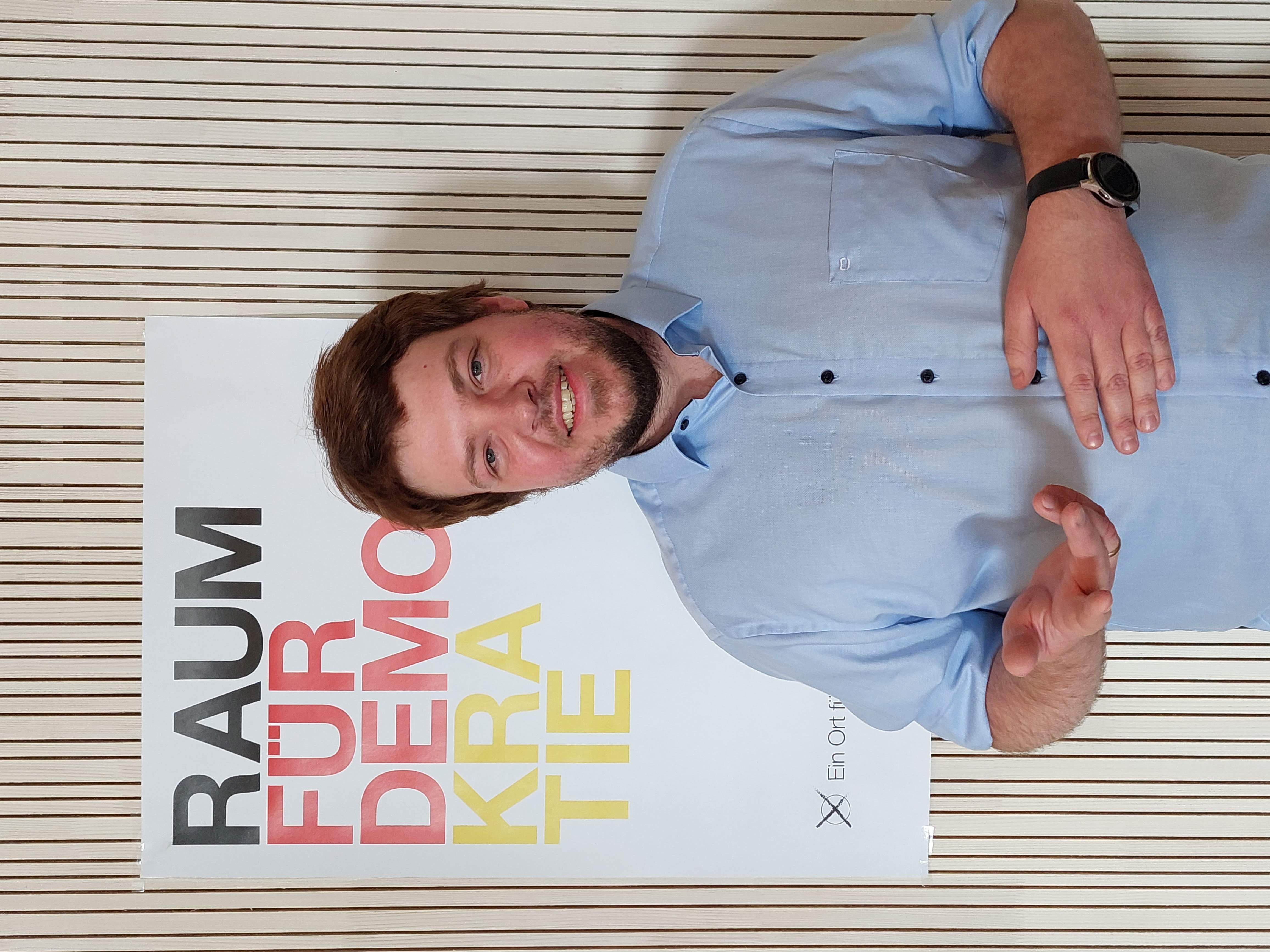 Ein lächelnder Mann im hellblauen Hemd gestikuliert vor einem Poster mit der Aufschrift "RAUM FÜR DEMOKRATIE".