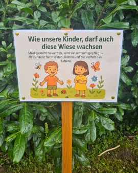 Ein Schild mit zwei gezeichneten Kindern steht vor einem grünen Busch.