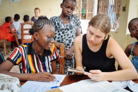 Eine Lernsituation: Ein Mädchen in Benin lernt und wird dabei von einer jungen Frau, die einen Freiwilligendienst als Don Bosco Volunteers macht, unterstützt. Um sie herum stehen zwei weitere Kinder, im Hintergrund sieht man eine andere Lerngruppe.