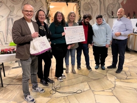 Zwei Männer, drei Frauen und zwei junge Männer stehen in einer Reihe. Die Frauen halten einen großen Scheck in der Hand.