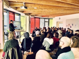 In einem runden Raum mit bunten Glasfenstern und Holzdecke sitzen Menschen und blicken nach vorne zu einem Mann, der neben einem Bildschirm steht.