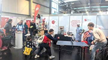 Auf einer Messe stehen Menschen an einem Tresen, in der rechten Bildhälfte spielen zwei Jugendliche Tischtennis. Im Hintergrund steht ein Mann mit Kochschürze.