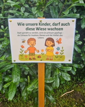 Ein Schild mit zwei gezeichneten Kindern steht vor einem grünen Busch.