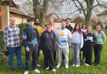 Elf junge und ältere Menschen stehen in einem Garten auf einer Wiese. Im Hintergrund sind zwei Bäume mit Herbstlaub zu sehen.
