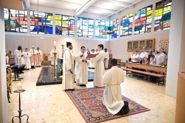 Emmanuel Eze kniet in einer Kutte gekleidet auf einem Teppich. Vor ihm steht ein Priester und liest aus einem Buch vor. Zwei Ministranten und weitere zehn Gäste stehen in der Kirche.