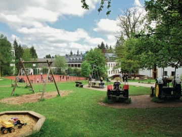 Spielplatz und Schulanlage mit Kindern, Traktoren und Sportplatz an einem sonnigen Tag.
